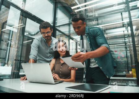Searching for solutions. Group of three young and positive employees using modern technologies and discussing something while working in modern office Stock Photo