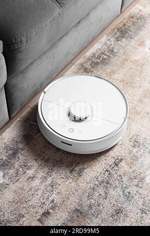 White robot vacuum cleaner on the carpet in the living room near the sofa. Cleaning the apartment with a modern smart vacuum cleaner. Stock Photo