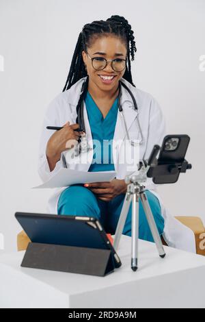 Happy african american doctor woman wears medical coat holding and using smartphone with digital tablet against white background Stock Photo