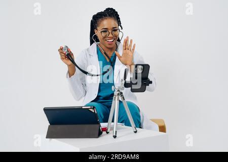 Happy african american doctor woman wears medical coat holding and using smartphone with digital tablet against white background Stock Photo