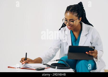 Smiling african american female doctor gp wears white medical coat using tablet at workplace gives remote online consultation, telemedicine chat Stock Photo