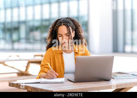 Focused trendy hispanic or brazilian curly haired girl, student or freelancer, sitting outdoors with a laptop, working or studying online, concentrated writing down information in her notepad Stock Photo