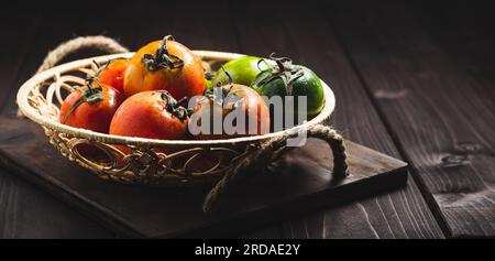 Fresh colorful ripe fall or summer heirloom variety tomatoes in a basket on dark wooden background. Autumn and harvest concept. Still life, close up Stock Photo