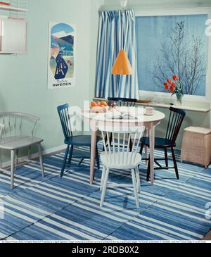 Kitchen in the past. A kitchen with a round kitchen table with four kitchen chairs. On the floor are home-woven rag rugs in blue and white fabric. A yellow tin kitchen lamp hangs over the table. A classic String shelf hangs on the wall. Sweden 1958. Conard ref BV98-6 Stock Photo