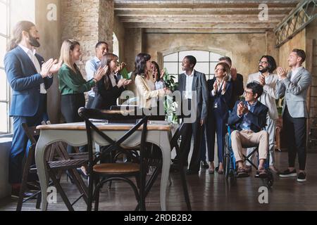 Diverse work team celebrates success in a modern rustic office. Middle-aged Japanese woman congratulates young African woman, as others applaud. Stock Photo