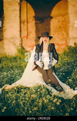 Woman blonde in hat sitting on chair in nature on background of old building with arches. Lady is holding an aquarium and a mannequin head in her hand Stock Photo