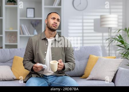 African American young man suffering from illness, flu, infection, runny nose at home. Sitting on the sofa and taking medicine, holding a cup with a drink and a napkin. Stock Photo