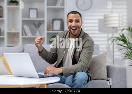 Portrait of a happy young African American man sitting on the couch at home and using a laptop. With the gesture of the winner, he rejoices at the camera. Stock Photo