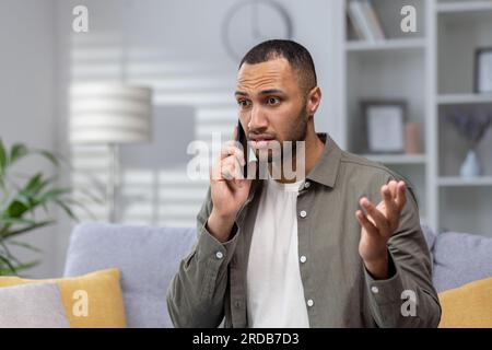 Portrait of upset young hispanic man talking on the phone at home. Worriedly spreads his hands, solves problems on the phone, received unpleasant news. Stock Photo