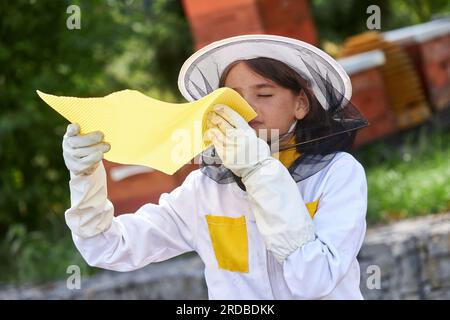 Girl smelling honeycomb sheet with eyes closed wearing protective suit at apiary Stock Photo