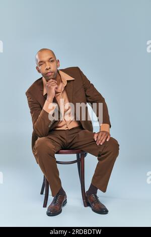 real people, full length of african american man with myasthenia gravis sitting on wooden chair on grey background, dark skinned person in suit lookin Stock Photo