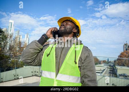 young male construction worker of african ethnicity wearing safety clothes and hardhat standing outdoors talking on the phone discussing with his empl Stock Photo