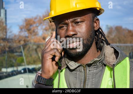 closeup of young african ethnicity man civil engineer, employee at construction site, disappointed talking on the phone with his boss, listening atten Stock Photo