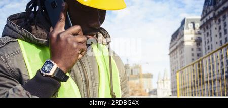 closeup banner of black male civil engineer wearing yellow safety helmet, outside at construction site talking on phone listening attentively, copy sp Stock Photo