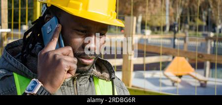 closeup banner of black male civil engineer wearing yellow safety helmet, outside at construction site talking on phone listening attentively, copy sp Stock Photo