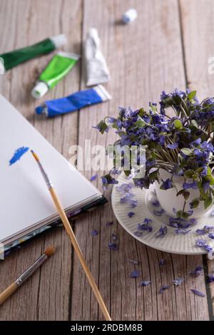 photo of a wilted Hepatica standing in a white cup next to a white sheet of paper, paints, paint brushes on the table Stock Photo