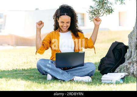 Cheerful curly mixed race female student or freelancer, sits outdoors on the grass with a laptop, rejoicing in success, passed the exam, finished the project, gesturing with both hands, smile happily Stock Photo