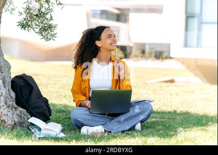 Happy smart pretty mixed race curly haired young woman, successful student or freelancer, sit outdoors on a grass with laptop, working or studying online, dreamily looks away, ponders an idea, smile Stock Photo