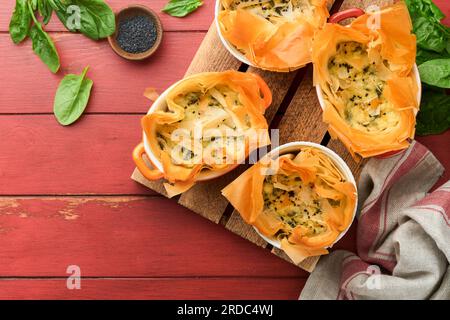 Filo pies with soft feta cheese and spinach in ceramic molds on old red wooden table backgrounds. Filo portions pies. Small Baked Spanakopita pies. To Stock Photo