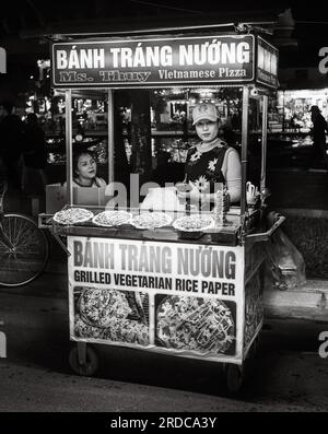 A Vietnamese woman holds her mobile phone and stands at her street food stall at night selling Banh Trang Nuong, or Vietnamese pizza, in Hoi An, Vietn Stock Photo