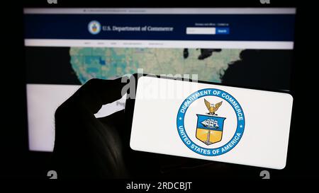 Person holding cellphone with seal of the United States Department of Commerce (DOC) on screen in front of webpage. Focus on phone display. Stock Photo