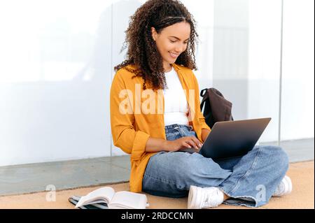 Using laptop. Education concept, female student. Lovely stylish curly haired mixed race girl, sit near the university campus, using laptop, preparing for the exam, doing homework, project task, smile Stock Photo