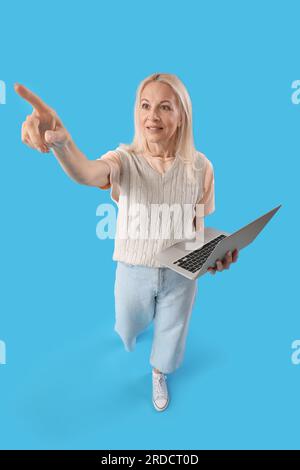 Mature female programmer with laptop pointing at something on blue background Stock Photo