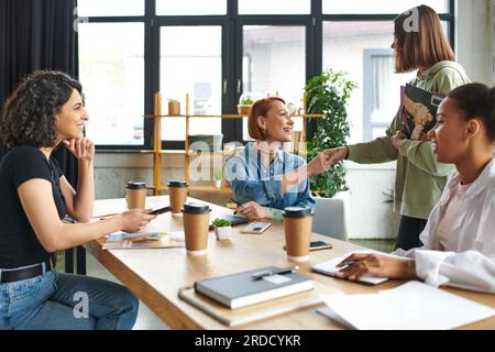 young woman holding magazines and shaking hands with motivation coach while meeting with friendly female community near takeaway drinks in interest cl Stock Photo