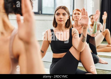young, concentrated and tattooed woman in sportswear meditating in lord of fishes pose during yoga class with multiethnic friends, wellness and mental Stock Photo