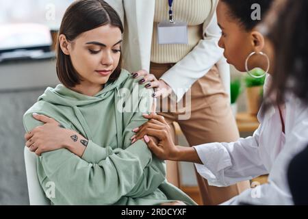 professional psychologist and african american girlfriend touching depressed and offended tattooed woman sitting with folded arms during supportive th Stock Photo