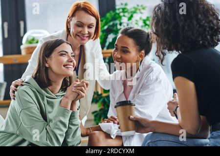 inspired and excited woman sitting and talking to multiethnic female friends and smiling psychologist during supportive session in consulting room, mo Stock Photo