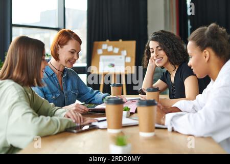 cheerful redhead woman reading magazine to smiling multiethnic female friends while spending positive time with takeaway drinks in women interest club Stock Photo