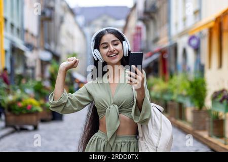 Young beautiful id woman walks in the city, tourist holds phone in hands, uses headphones to listen to music and online radio podcasts, dances and sings along happily. Stock Photo