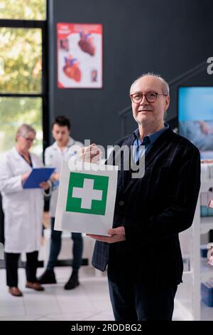 Senior man holding purchase shopping bag in drugstore portrait. Pharmacy elderly smiling customer buying prescription medicaments, standing in apothecary and looking at camera Stock Photo
