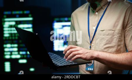 Precise mechaninc doing routine system scan, typing data on laptop of needed repairs in blade servers housing advanced data storage equipment supporting important IT workloads Stock Photo