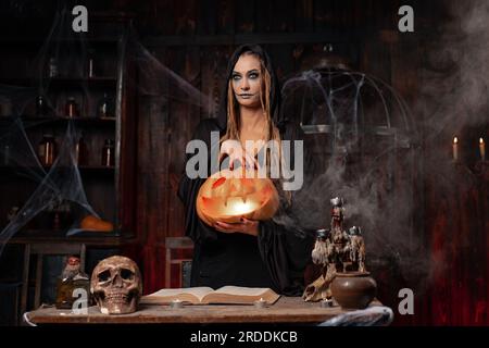 Halloween concept. Black witch holding Halloween pumpkin in hand with carved smily face in hand standing dark room. Female necromancer, dungeon. Jack o lantern head with light inside Stock Photo