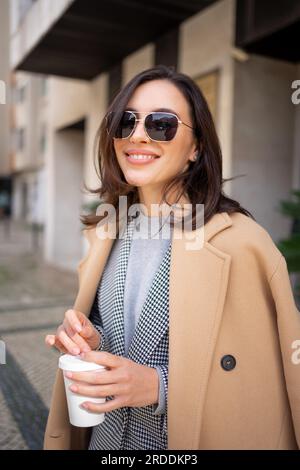 Streetstyle, street fashion concept: woman wearing trendy outfit walking in city. Cream trench coat, sunglasses. Looking camera, vertical photo Stock Photo