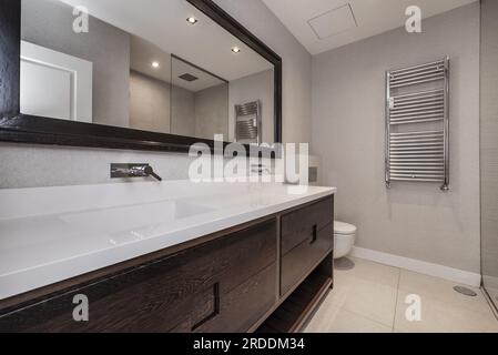 Bathroom with dark wood cabinet with drawers, long double sink and matching long mirror with wood frame and chrome radiator on the wall Stock Photo