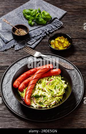 juicy cooked sausages with classic coleslaw salad in black bowl on dark wooden table with pickles and napkin at background, vertical view Stock Photo