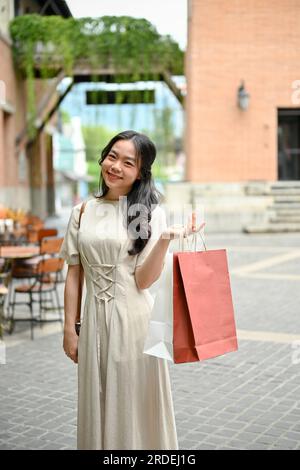 A portrait of a beautiful and happy young Asian woman in casual clothes walks with her shopping bags and enjoys shopping at a city square on the weeke Stock Photo