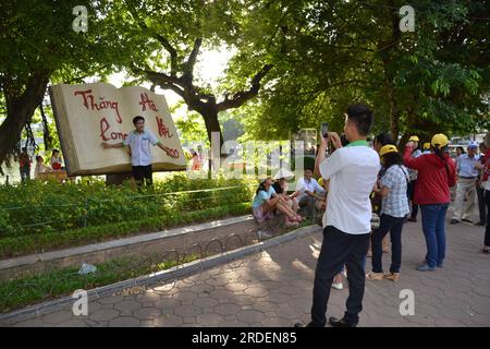 A young man takes a photo of a friend posing in front of the giant Open Book monument in Hoan Kiem Lake park in Hanoi, Vietnam, Asia; giant open book art sculpture celebrates 1000 or one thousand years of culture, founding of Hanoi Stock Photo