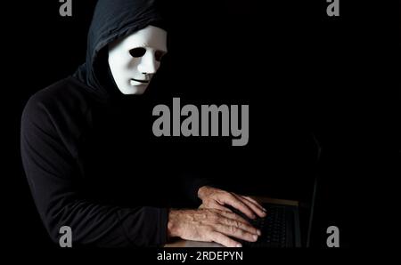 Male hacker dressed in black with a hood and a white mask using a computer Stock Photo