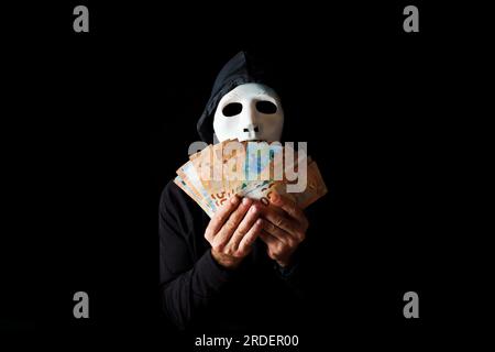 Hacker man dressed in black with white mask and black hood holding euro banknotes in his hands, cybercrime concept, isolated on black background Stock Photo