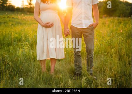 Couple holding hands in meadow during sunset & holding pregnant belly Stock Photo