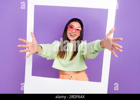 Photo of pleasant satisfied woman dressed green pullover stretching hands to you from photo frame isolated on purple color background Stock Photo