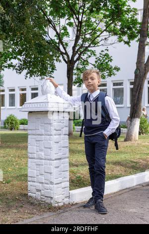 Happy boy in school uniform in a white shirt and blue vest with backpack stands near the school Stock Photo