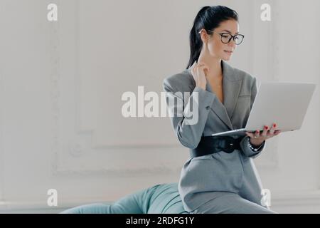 Serious elegant female administrator works from home, reading or watching a webinar on laptop, dressed in stylish business clothes, leaning on a sofa. Stock Photo