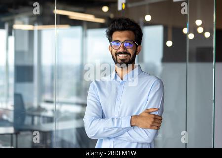 Portrait of a young Indian male designer, engineer, architect who is wearing glasses and a blue shirt smiling standing in the office and looking at the camera. Stock Photo