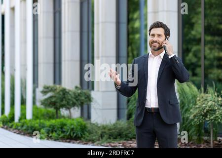 Mature successful boss talking on the phone, senior experienced businessman in business suit talking to clients using smartphone, man outside office building happy with achievement. Stock Photo