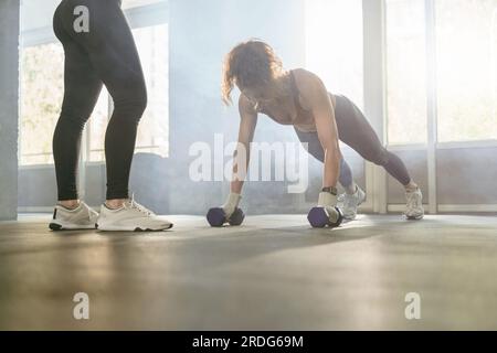 Young sporty woman doing push-ups with dumbbells while having workout with personal trainer in gym Stock Photo